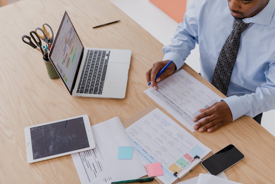 Overhead view of a professional at a desk with laptop, tablet, and papers — internal work and digital tools.